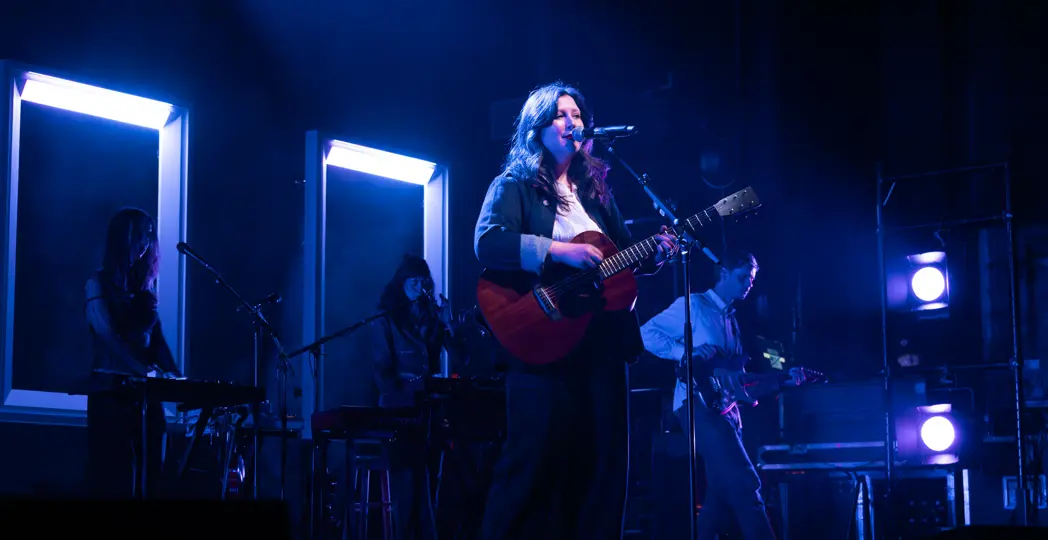 Lucy Dacus on a stage with blue lights, holding a guitar