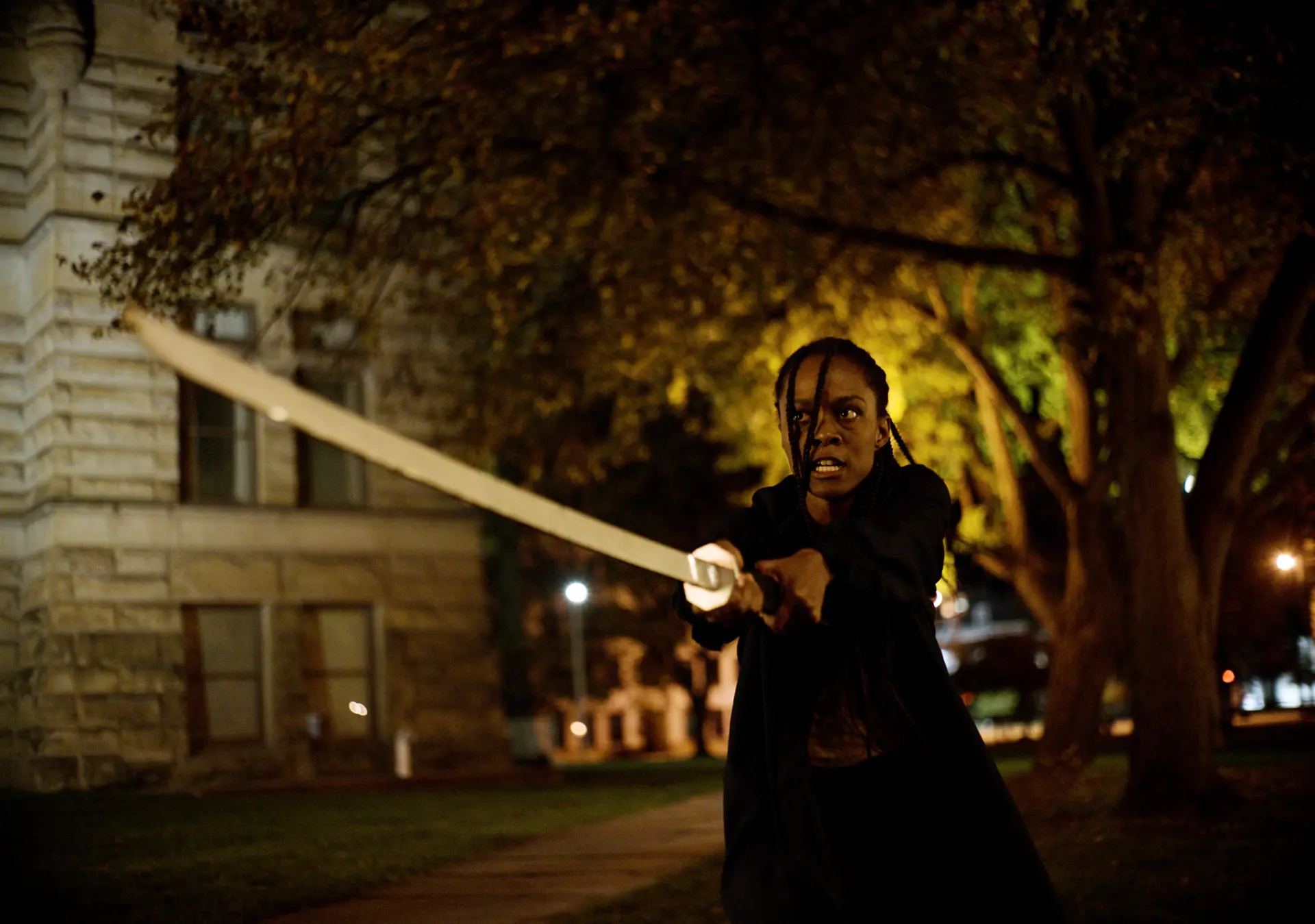 A woman holds a long metal sword in a nighttime scene.